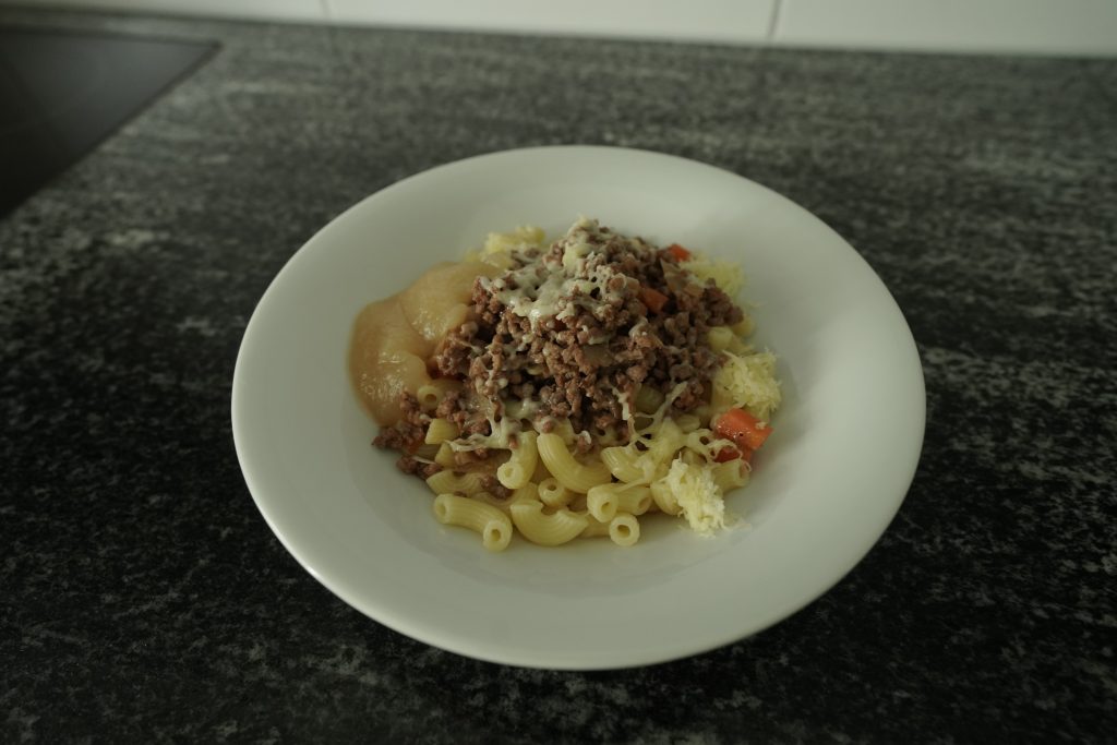 Pasta mit gehacktem Rindfleisch auf einem weissen Teller. Ebenfalls ist geriebener Käse und Apfelmus auf dem Teller.